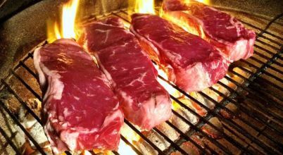 Thick steaks being cooked with direct heat on an open flam BBQ grill.